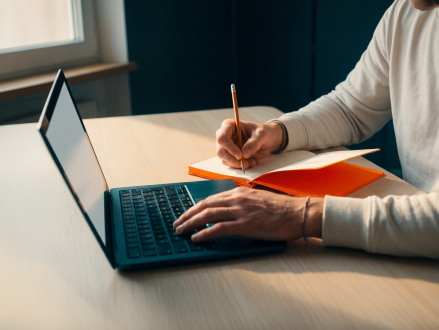 Person macht Notizen in einem Notizbuch neben einem geöffneten Laptop